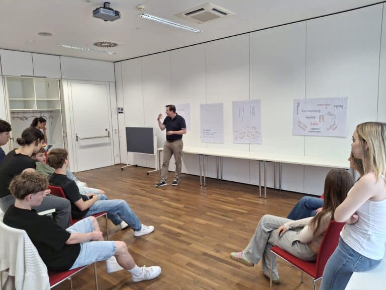 Vortragender erklärt einer Gruppe junger Leute in einem Seminarraum in Österreich etwas.